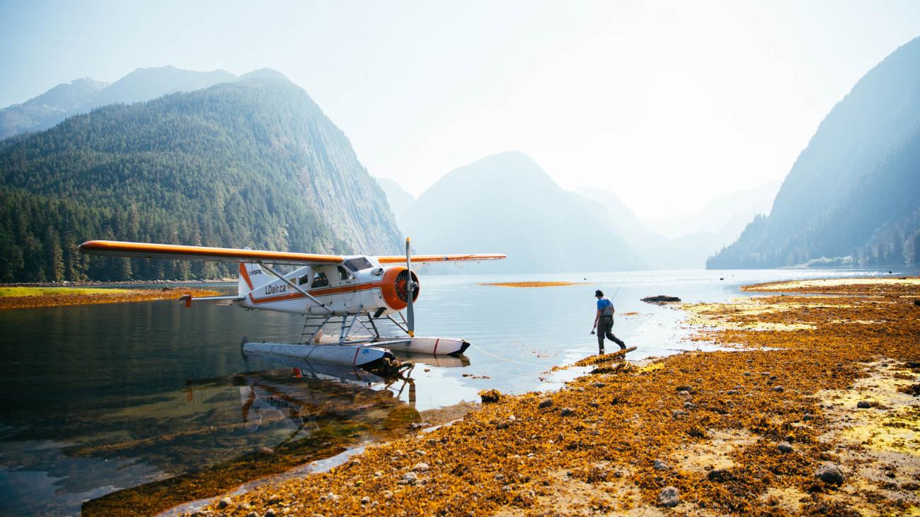 Seaplane parked on a serene lake, surrounded by mountains. There is a person walking on the shore.