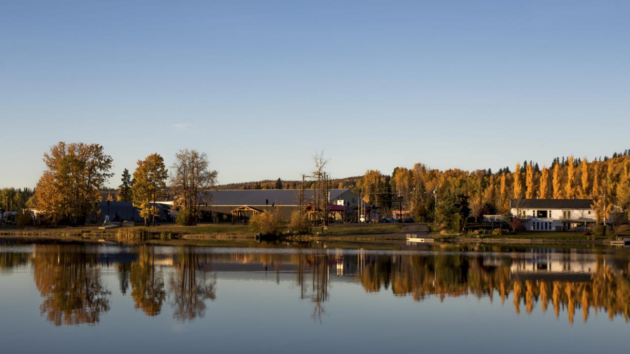 Autumn trees and village buildings reflected on a peaceful lake under a clear blue sky.