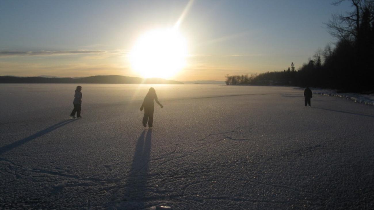 Group of people ice skating on frozen lake. The sun is setting in the background.