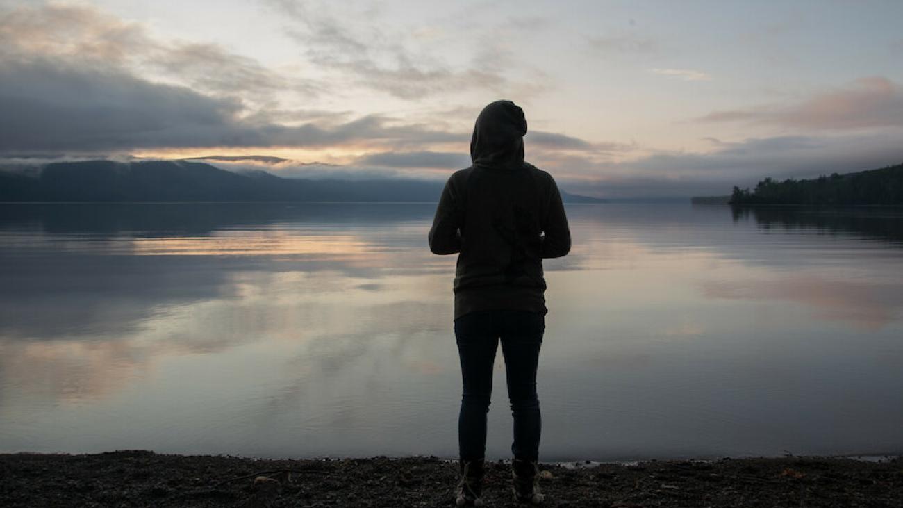 Person in a hoodie standing alone at the edge of a tranquil lake at dawn or dusk.