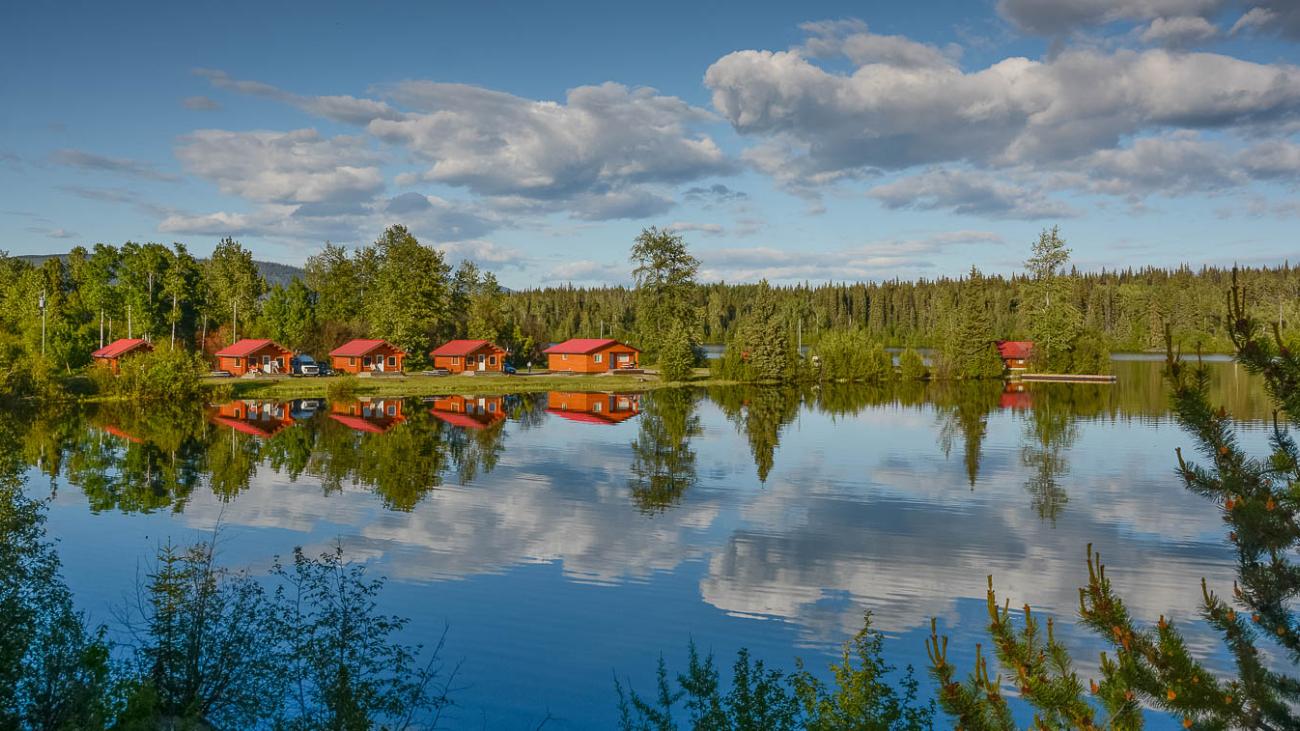Red cabins reflected in a calm lake with trees and clouds.