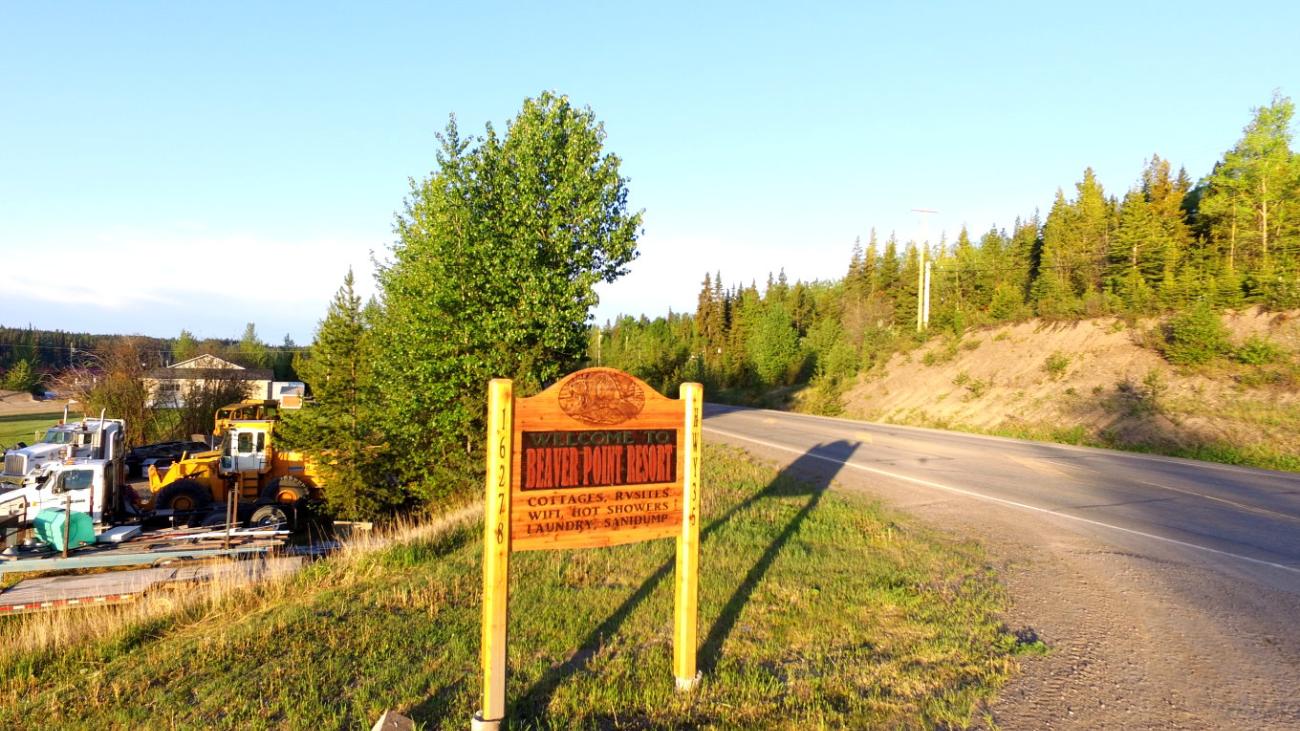 Dirt road with wooden signpost, surrounded by trees under a clear sky.