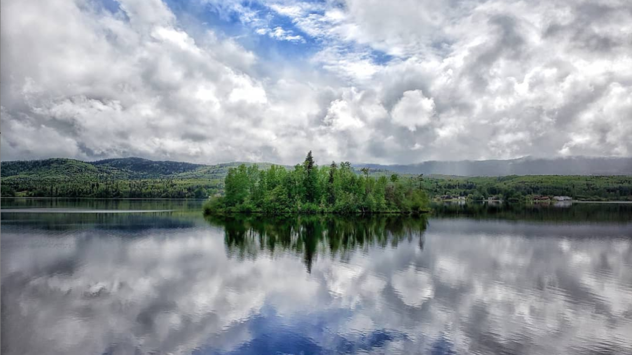 Small forested island reflected in a calm lake under a dramatic cloudy sky.
