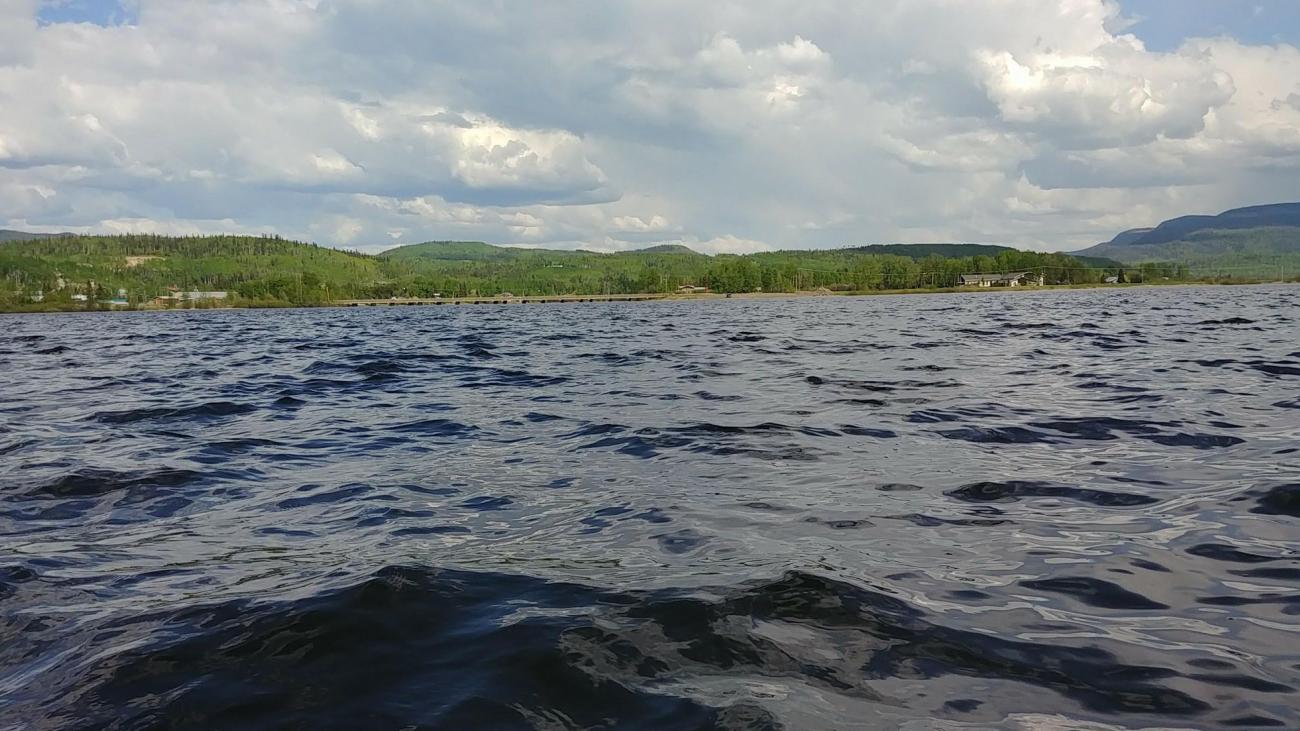 Gentle waves on a large lake with forested hills and cloudy skies in the background.