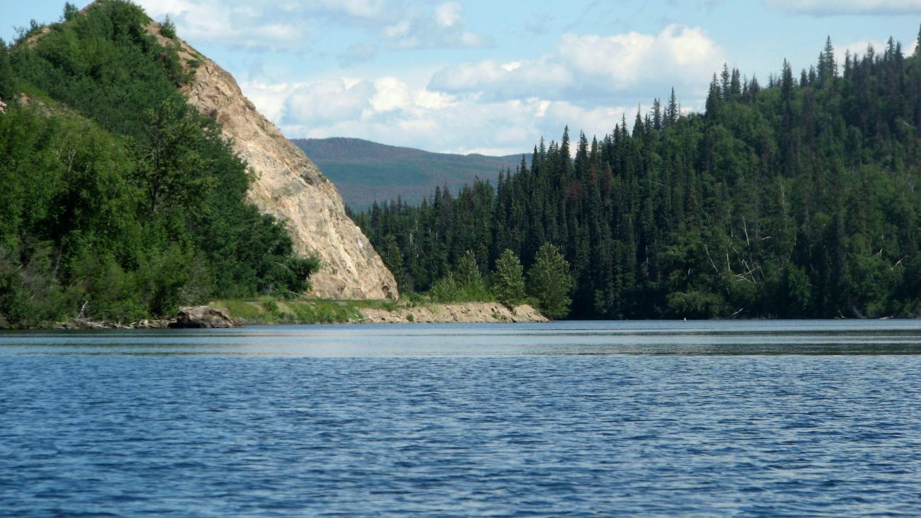 Blue lake bordered by evergreen trees and a rocky hill under a bright sky.