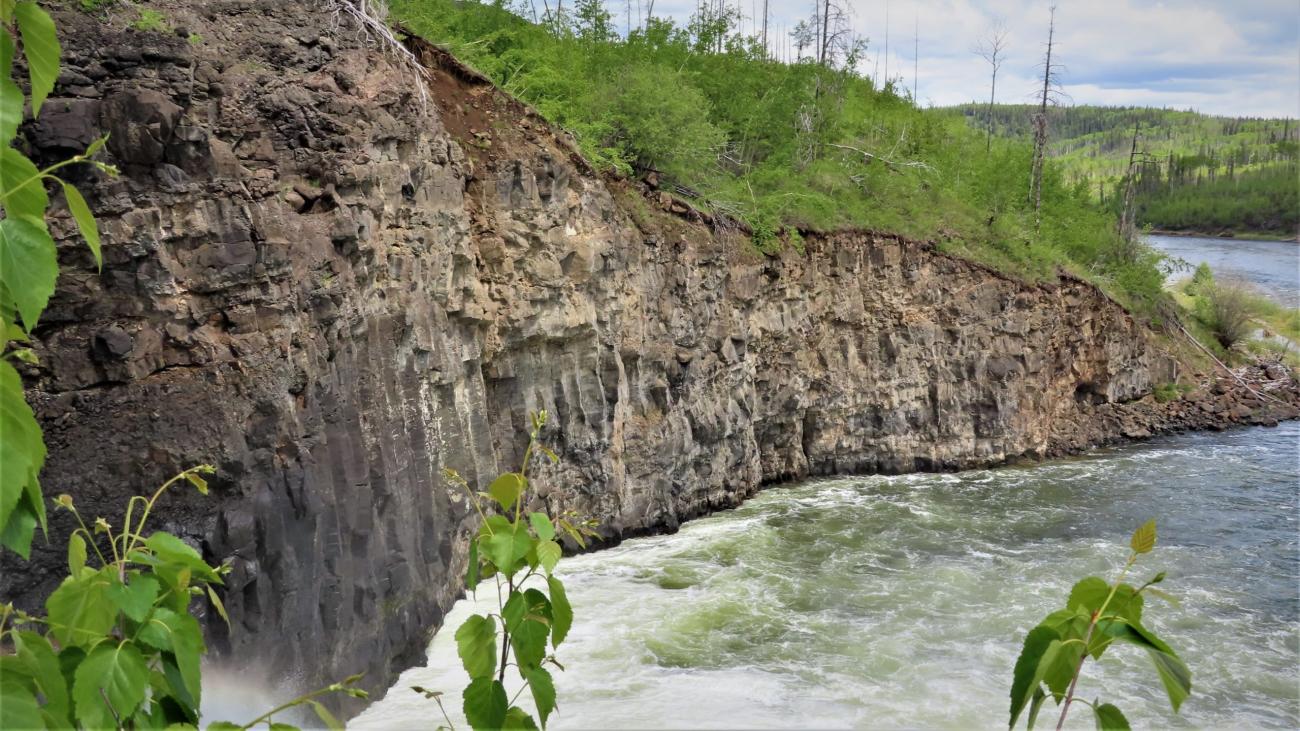 Steep rocky cliffside above a rushing river surrounded by lush greenery.