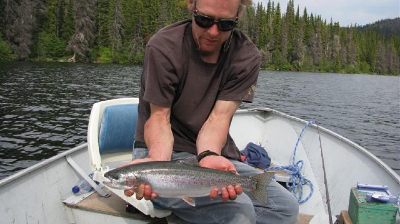 Man holding a fish on a boat in a lake surrounded by trees.