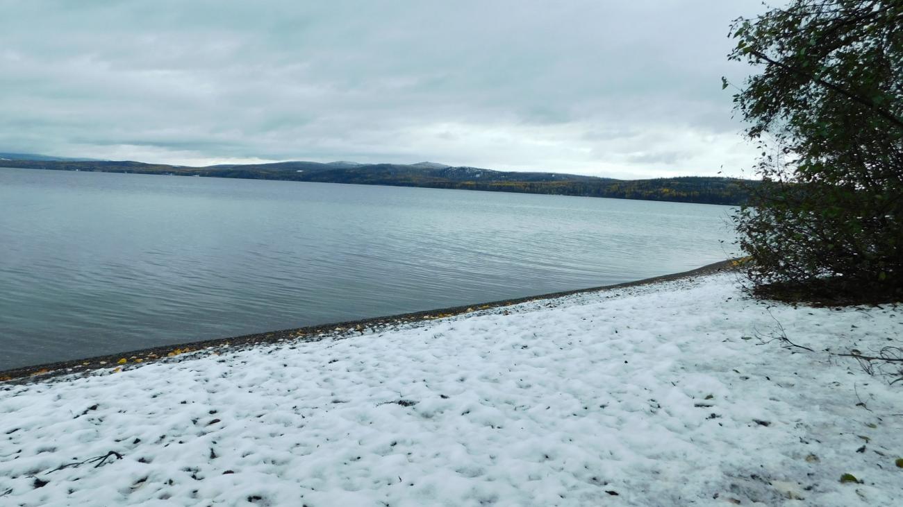 Snow-covered shoreline beside calm lake with distant forested hills.