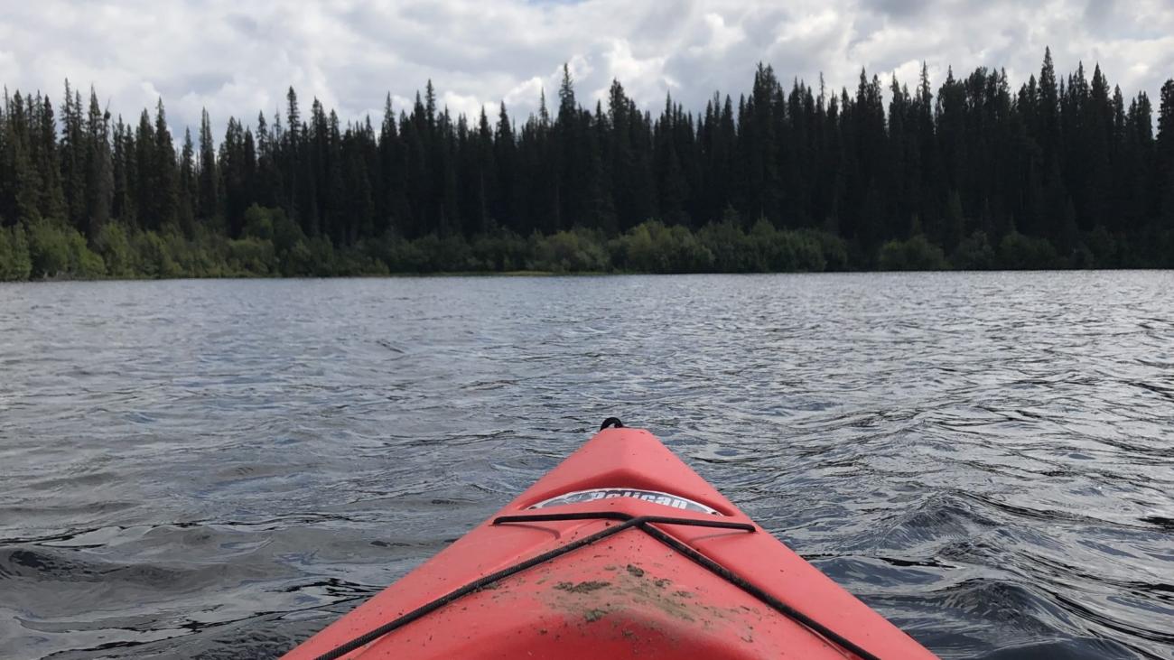 View from a red kayak on a calm forest lake under a partly cloudy sky.