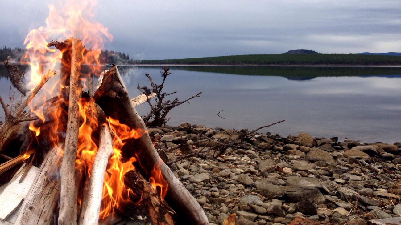 Lakeside campfire burning among driftwood on a rocky shore with still water in the background.