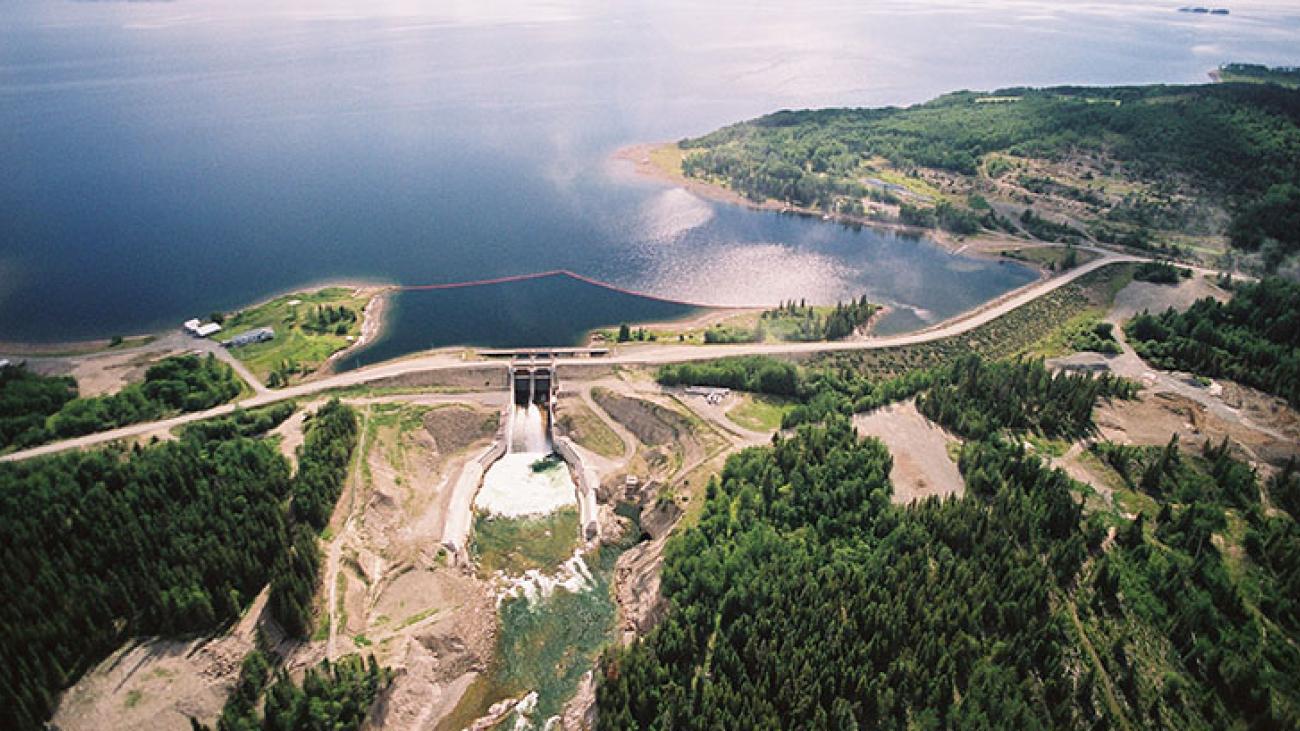 Aerial view of a dam and reservoir surrounded by forested hills and clear blue water.