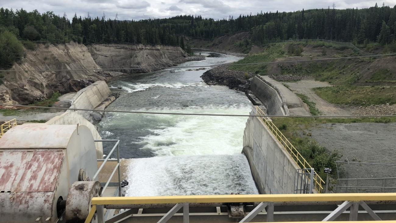 A dam releasing water into a forested river valley under cloudy skies.