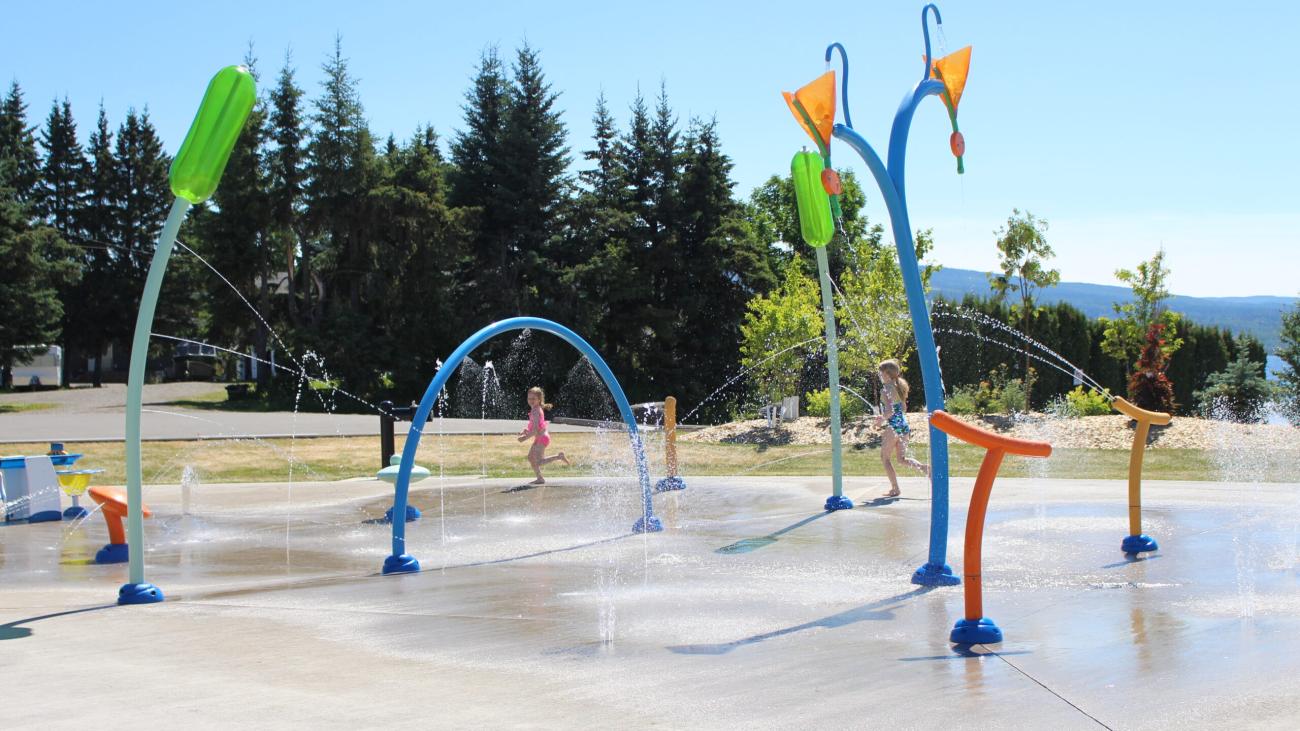 Children running through water jets at a splash park with mountains in the background.