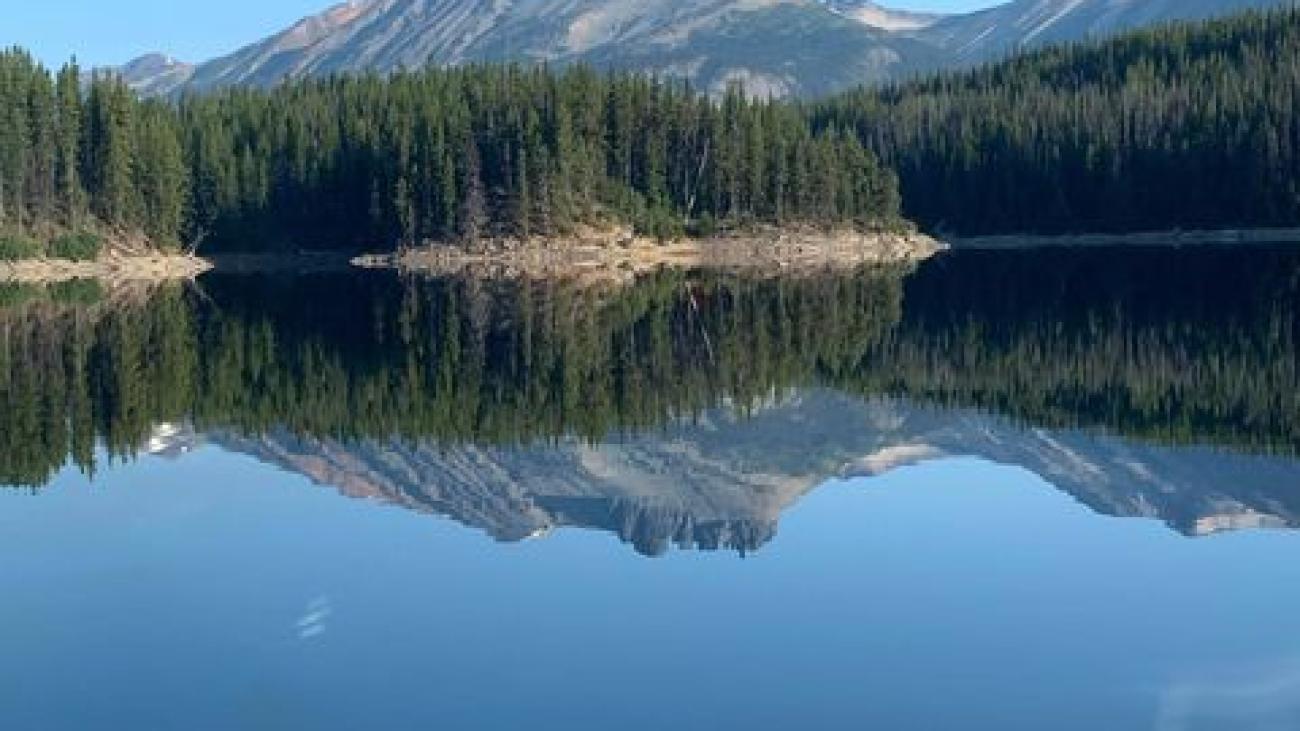 Mountain view from a boat on a glassy lake with pine forest and rugged peaks ahead.