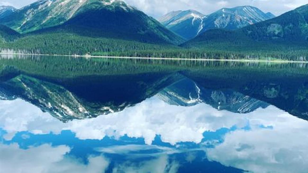 Snowy mountains mirrored perfectly in a still, clear lake under a partly cloudy sky.