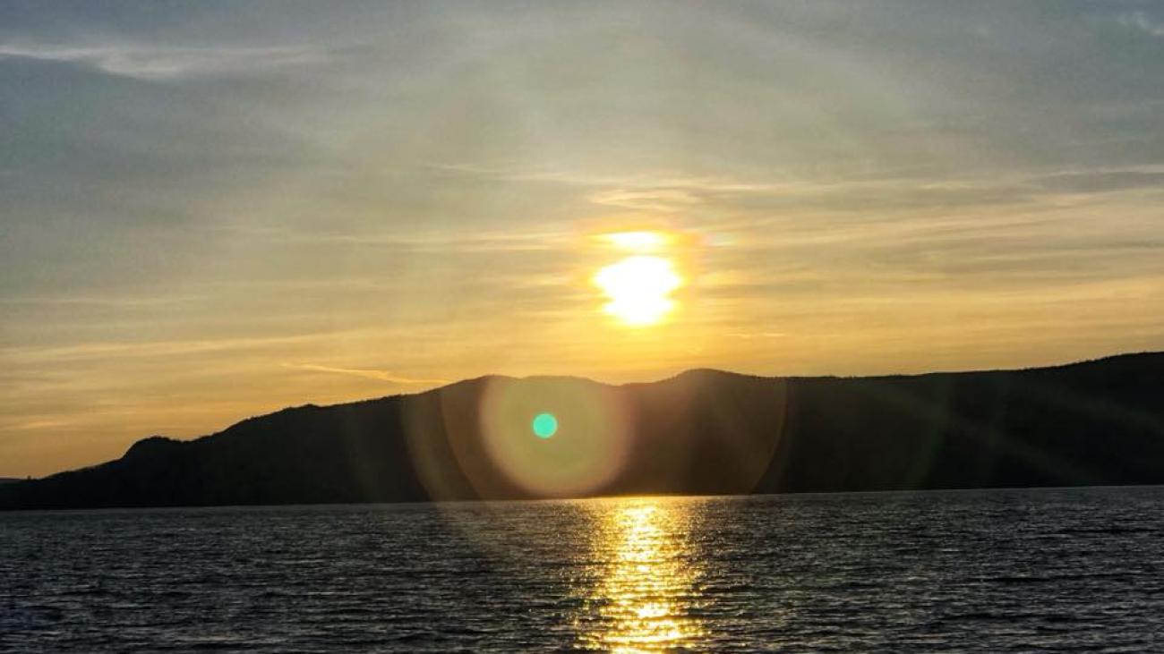 Bright sun setting over a calm lake with golden reflections and silhouetted hills.