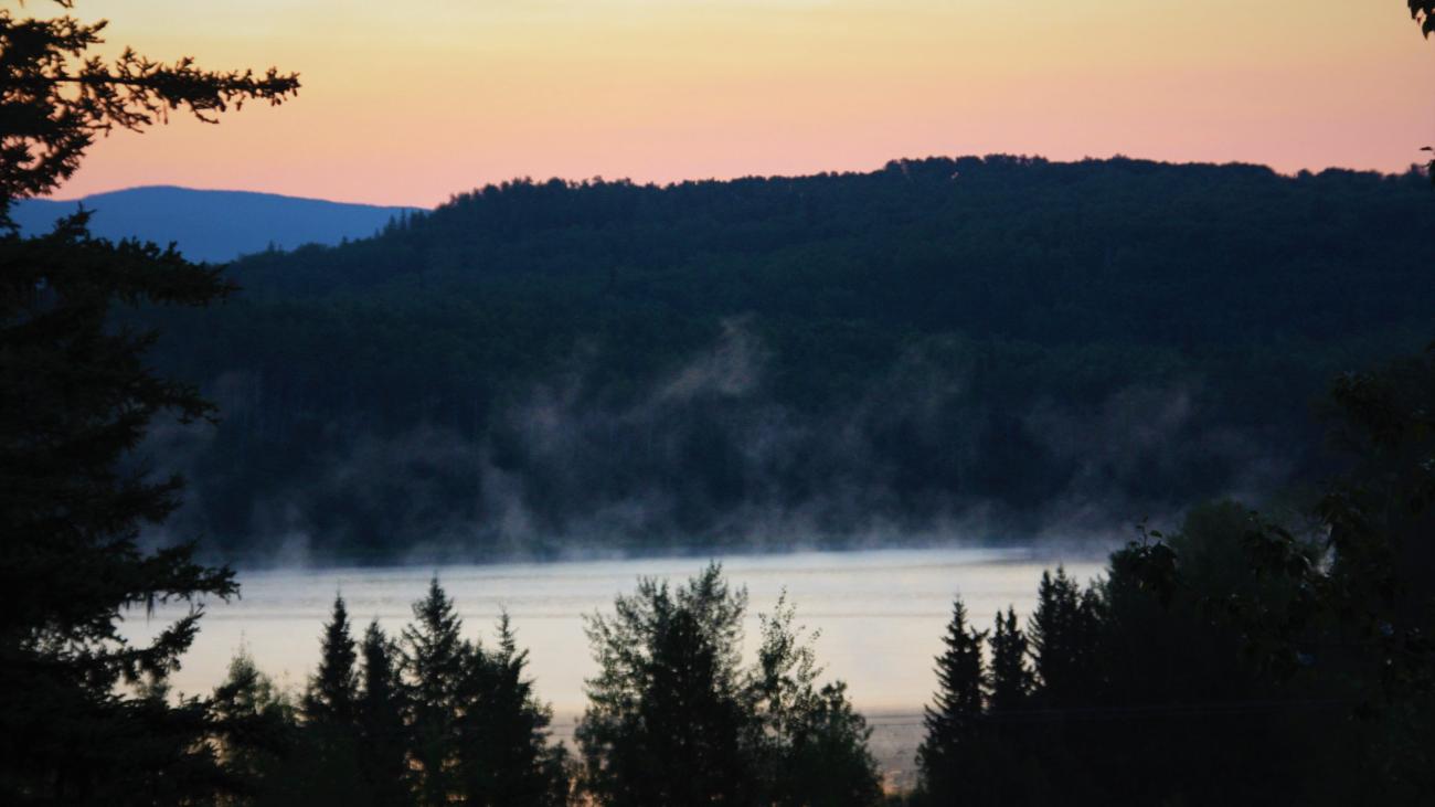 Mist rising from a tranquil lake at sunrise with forest-covered hills in the background.