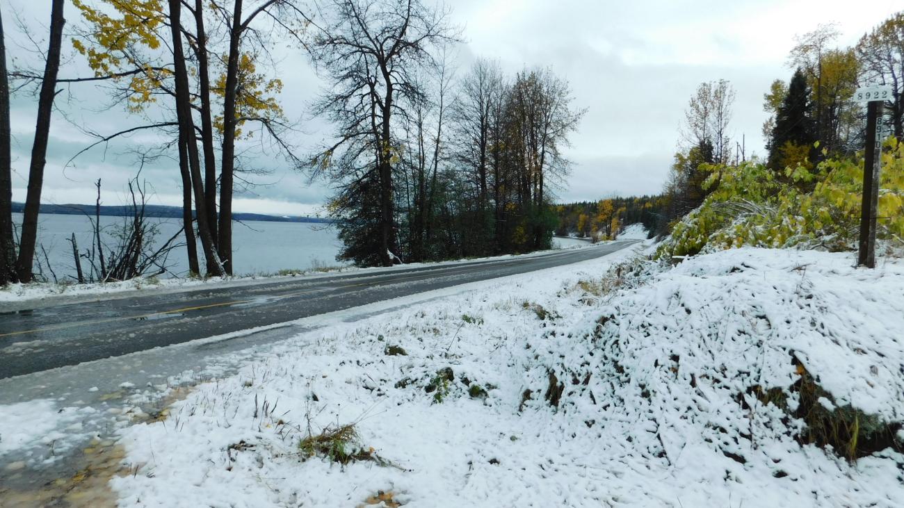 Early snowfall lining a lakeside road with autumn foliage and cloudy skies.