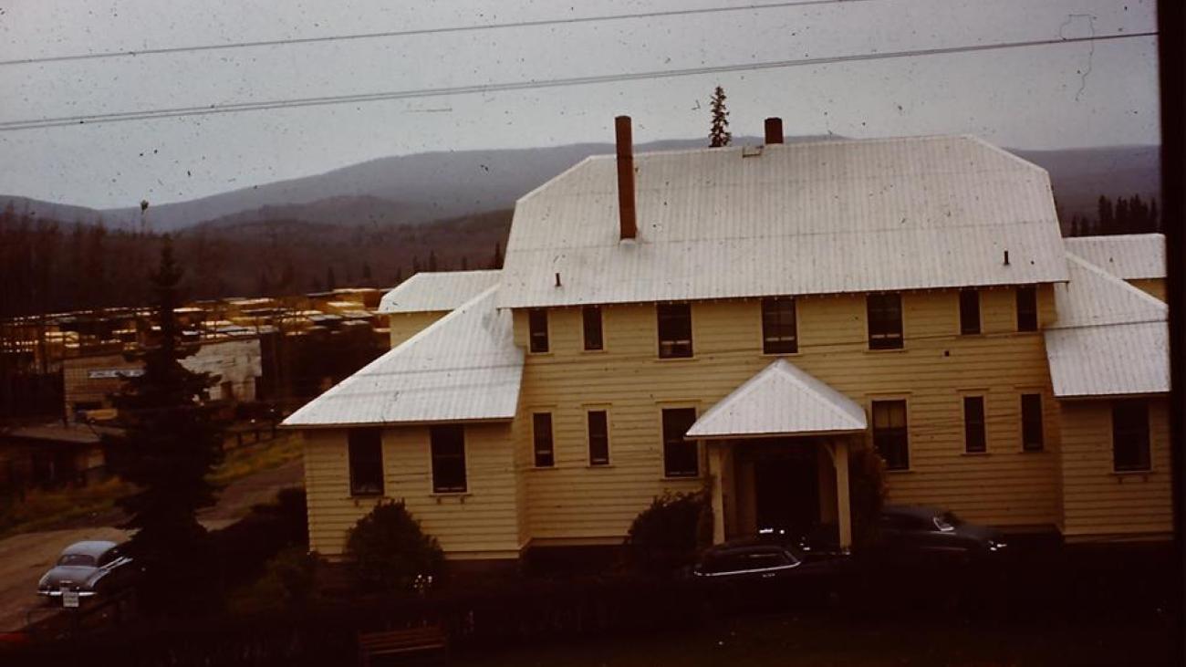 Historic two-story building with white roof, set against distant forested hills.