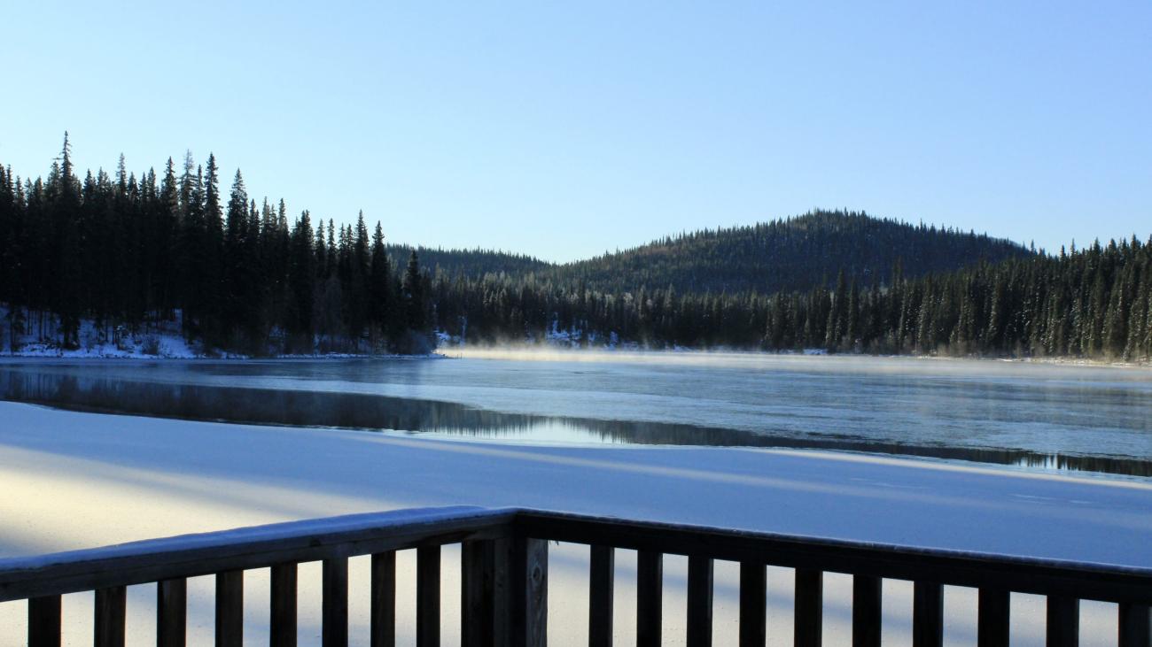 Snow-covered deck overlooks partially frozen lake surrounded by forested hills on a clear day.