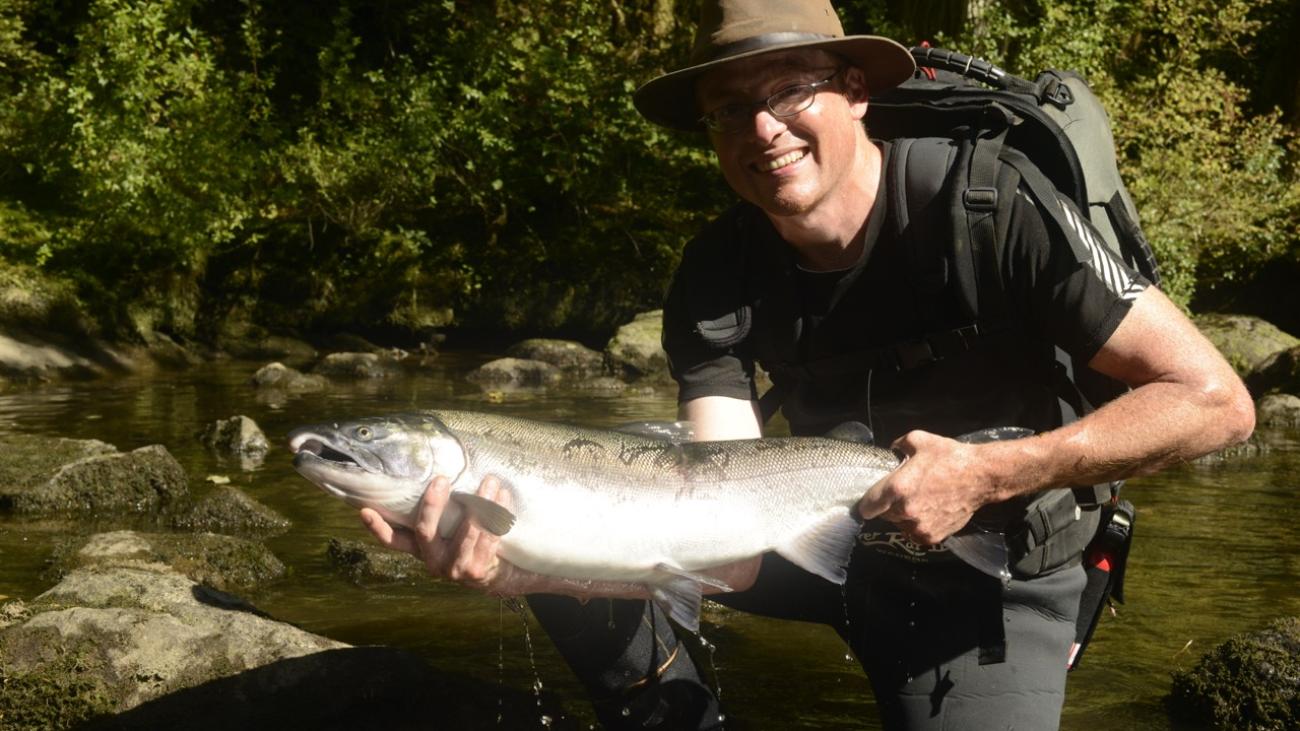 Man in hat holding a large fish in a forest stream.