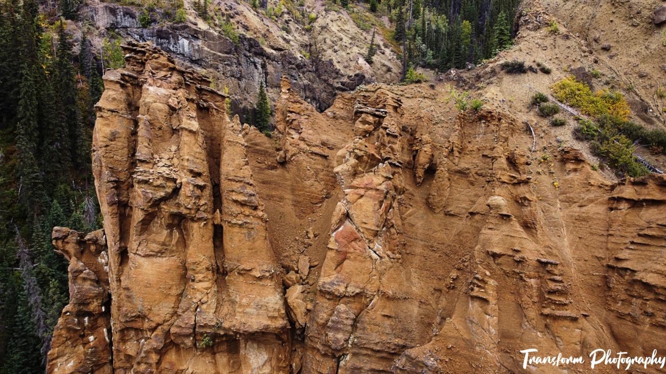 Steep, jagged red rock cliffs rise against a backdrop of sparse green trees and rugged terrain; "Transform Photography" watermark in the bottom right corner.