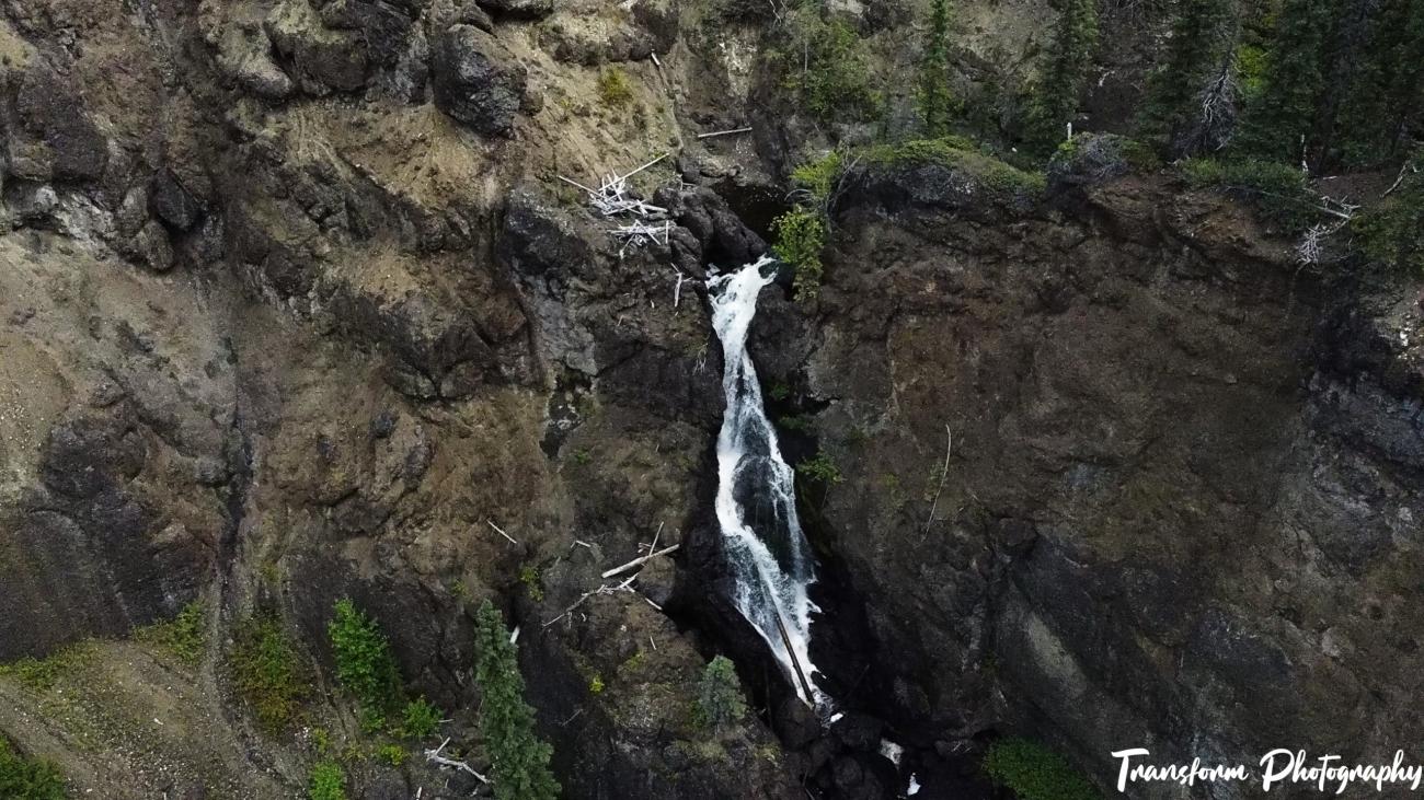 A narrow waterfall cascades down a rocky cliff surrounded by sparse trees and vegetation.