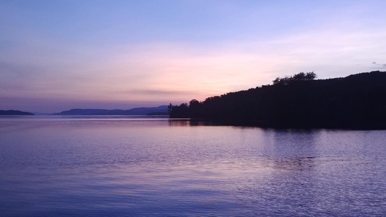 Soft purple and pink hues in a peaceful lakeside sunset over distant hills.