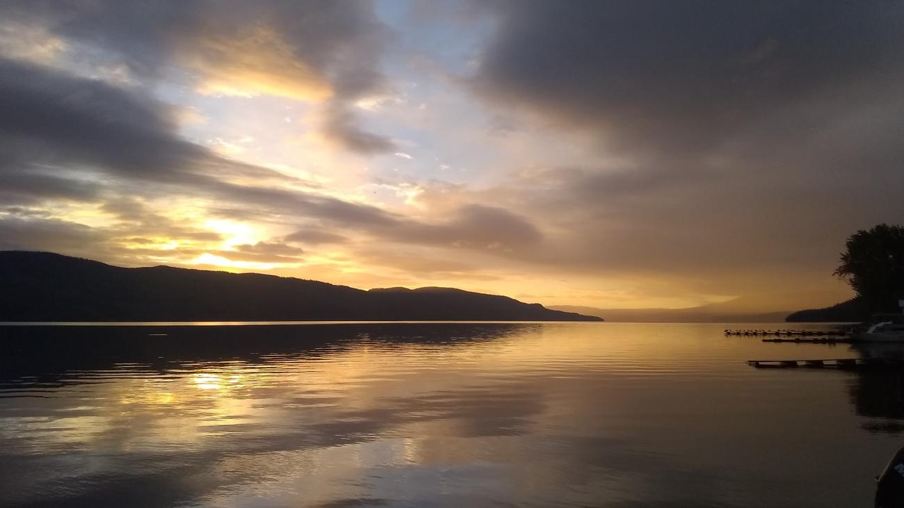 View from a wooden dock with boats, facing a golden lake sunset under dramatic clouds.