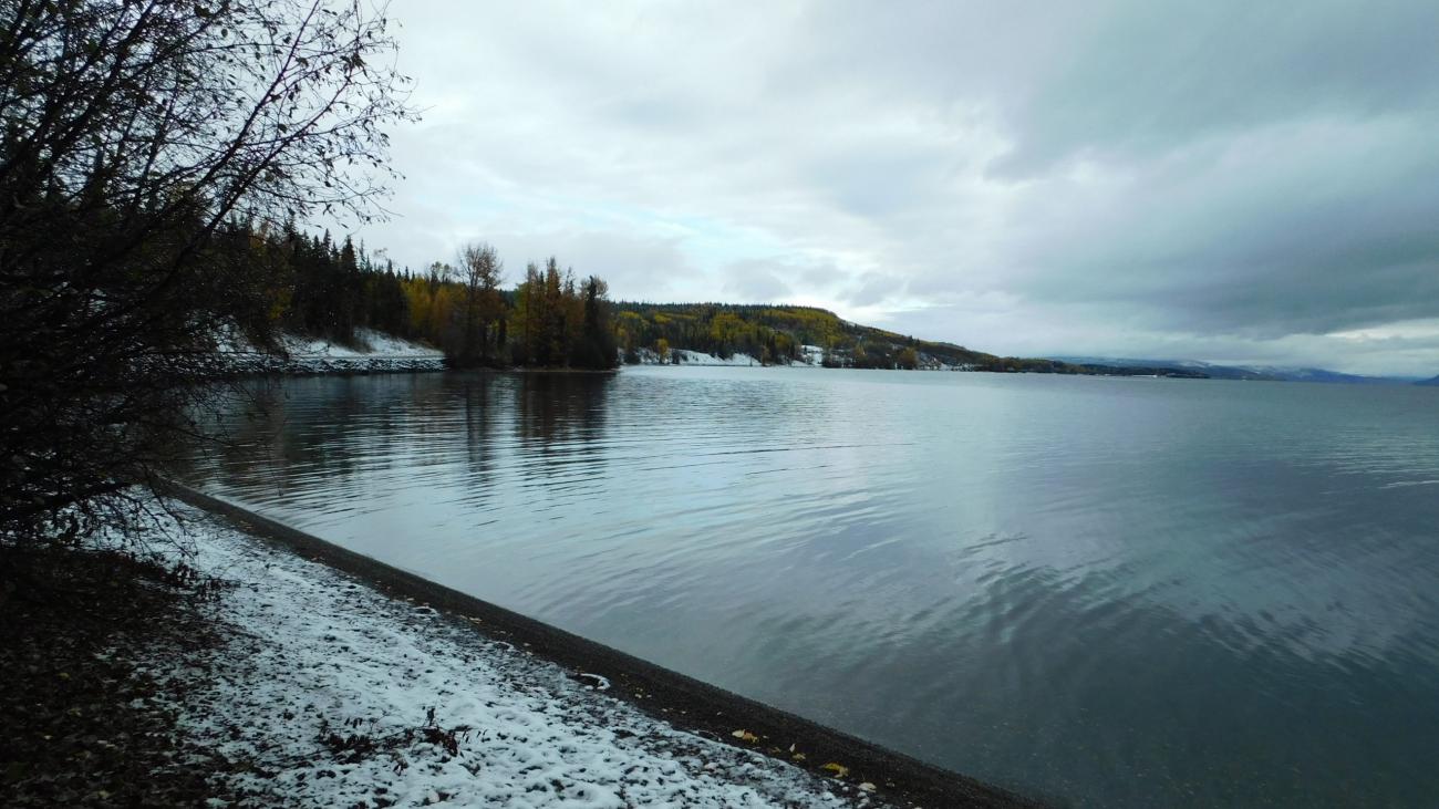 Snow-dusted shoreline meets calm lake beneath overcast autumn sky.