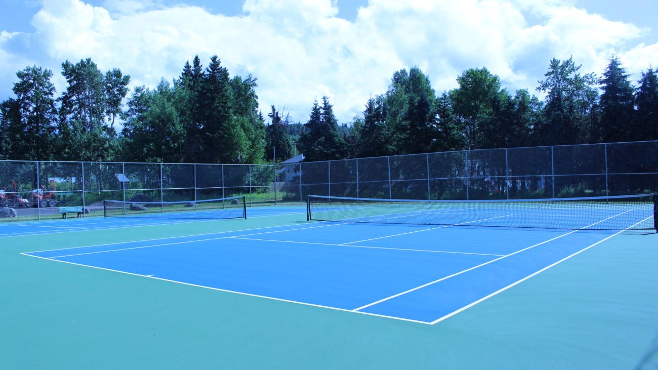 Bright blue tennis court surrounded by tall trees and chain-link fence under blue sky.