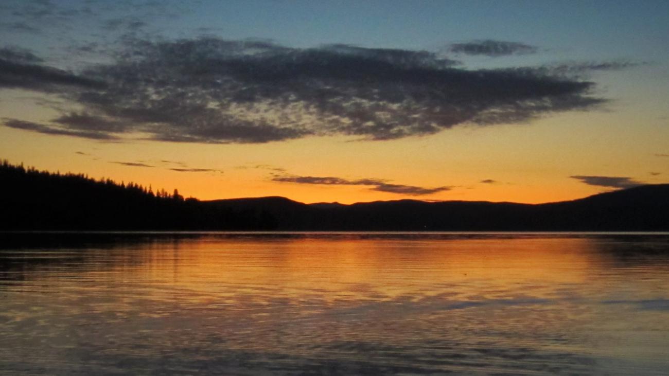Sunset over a calm lake with silhouetted trees and mountains.