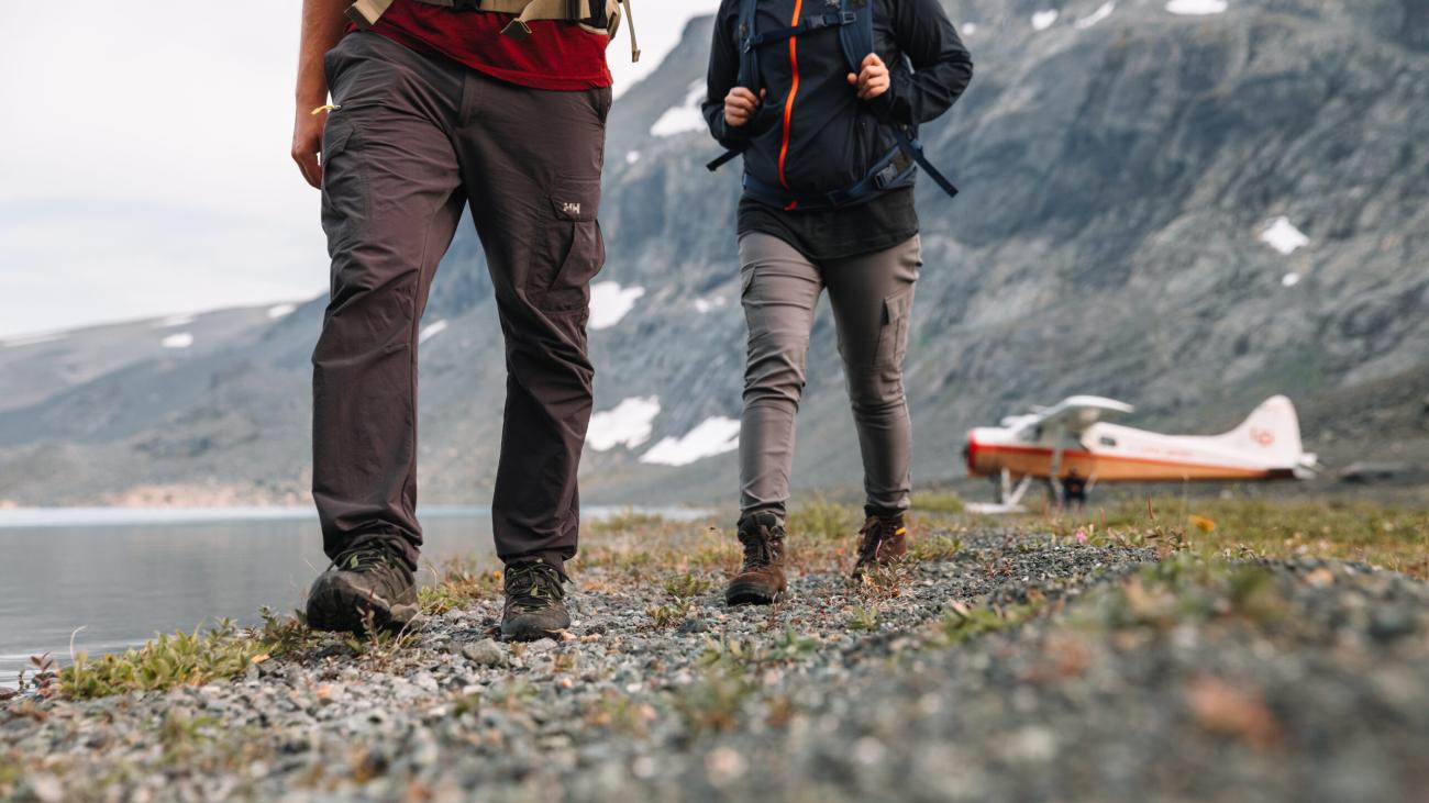 Two hikers walking on a rocky path by a lake. There is a seaplane parked in the background.