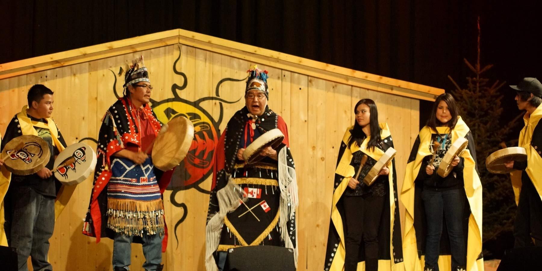 First Nations peoples in traditional attire playing drums on stage with wooden backdrop.